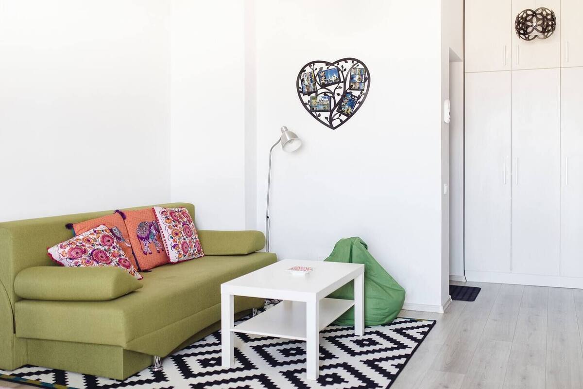 A room with a green sofa, a white table, a patterned rug, a heart-shaped photo decoration, and a wardrobe.