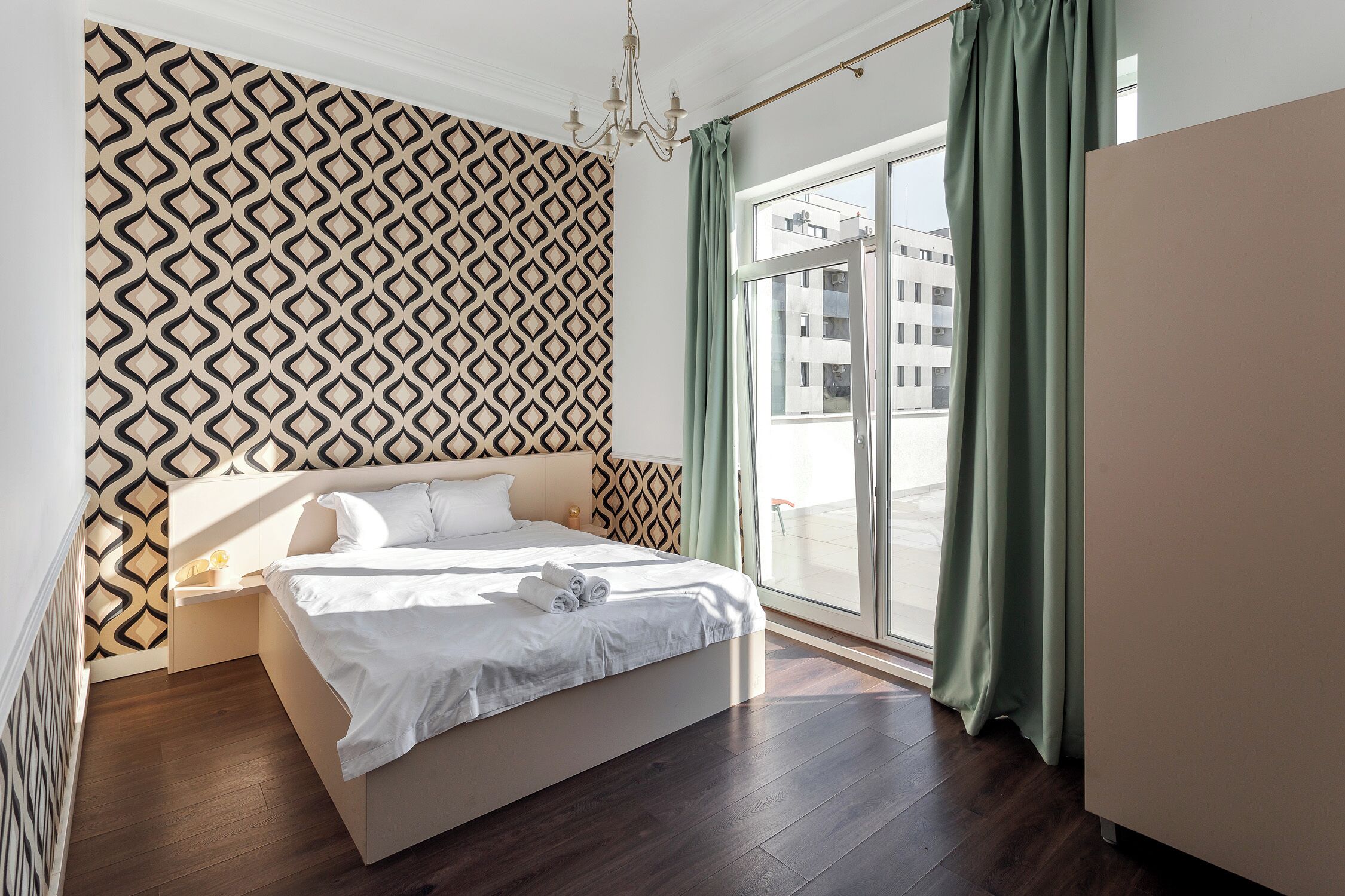Um quarto moderno com papel de parede estampado, uma cama grande, uma mesinha de cabeceira, uma cômoda e vista para a cidade através de uma janela ampla.