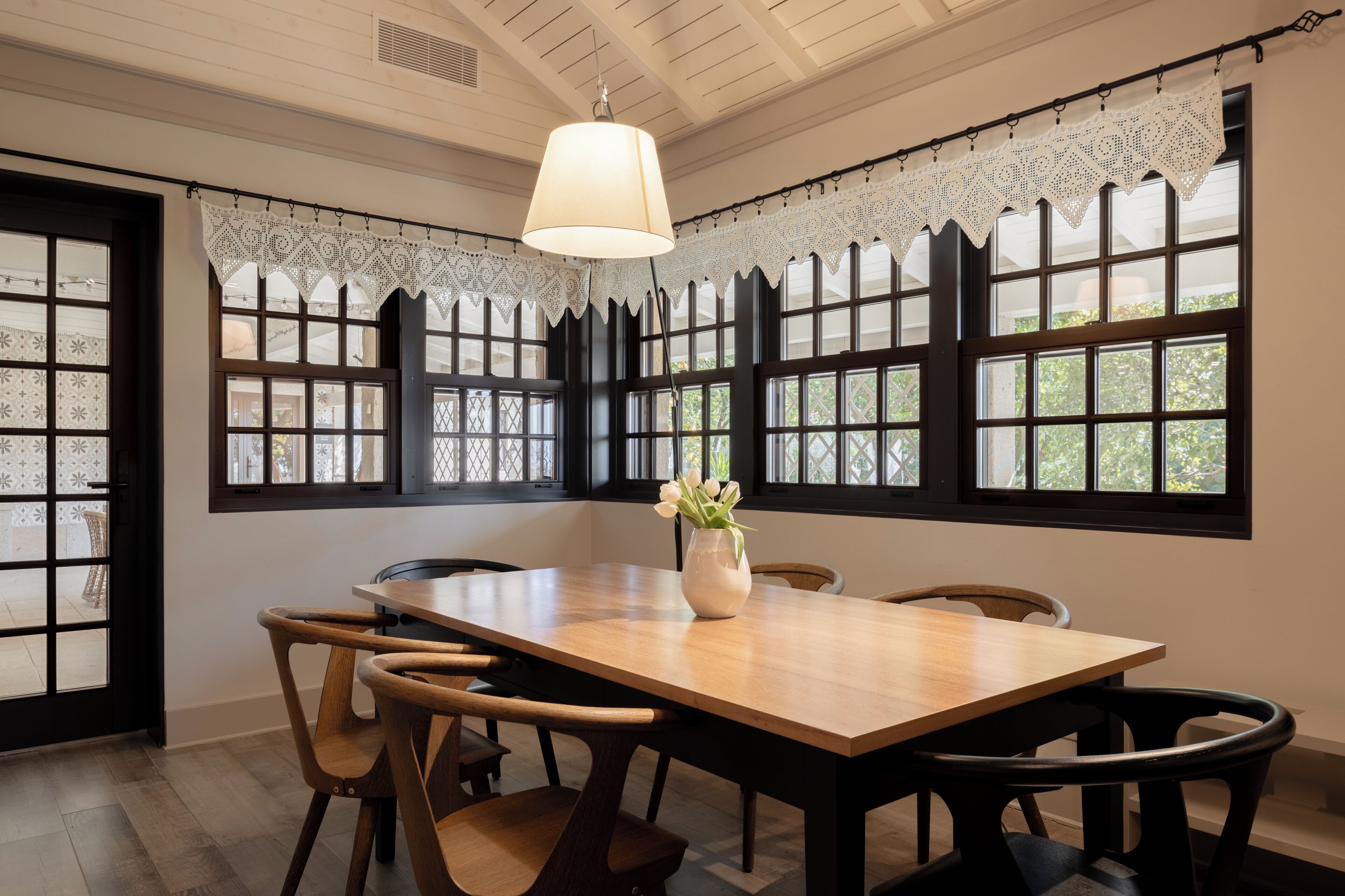 A dining area with a wooden table, chairs, lace curtains, and a hanging light fixture.