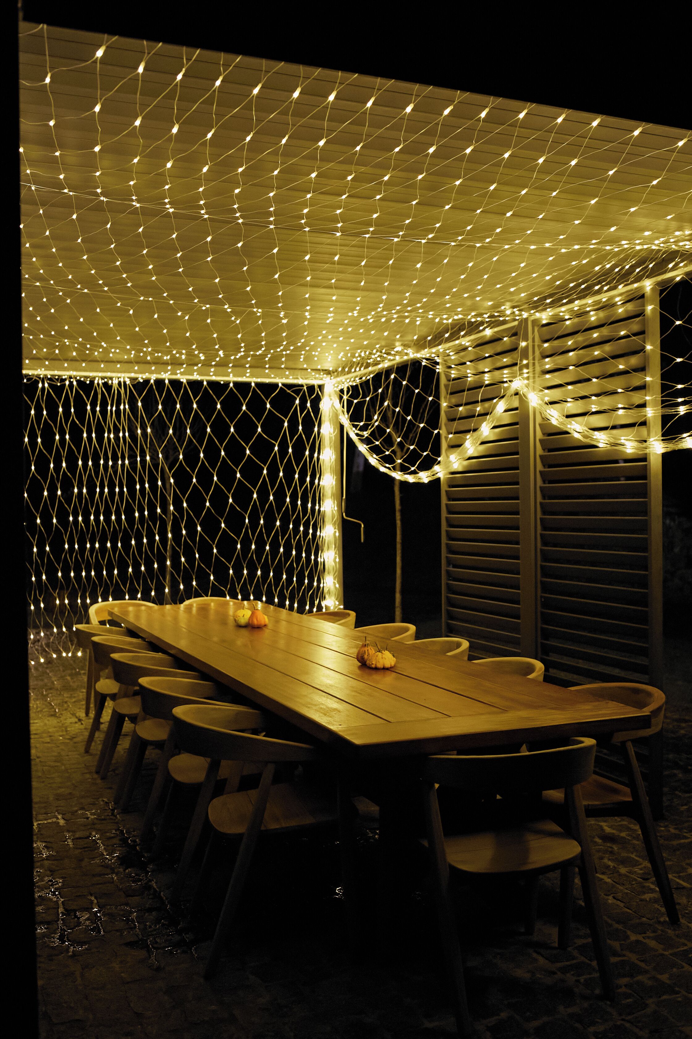 Un área de comedor al aire libre con una larga mesa de madera y sillas, rodeada por un recinto cubierto de mosquiteras y luces colgantes.