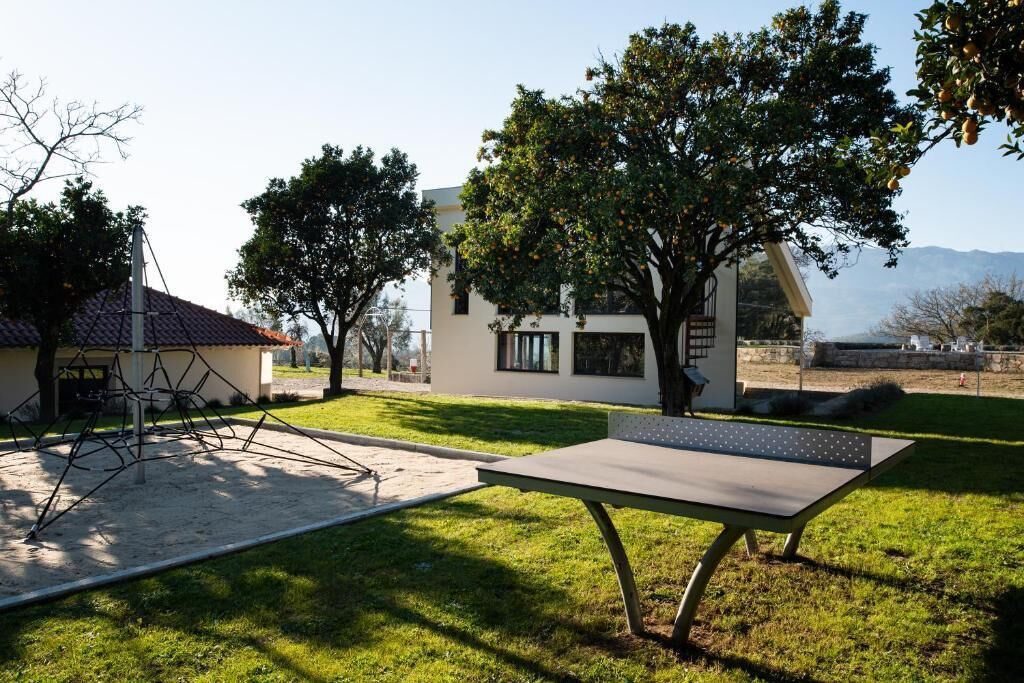 Un espacio exterior moderno con una mesa y sillas de metal, rodeado de árboles y un edificio con techo inclinado.