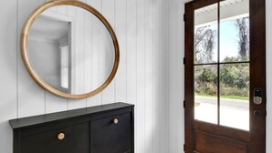 A dark wooden cabinet with two drawers and a round mirror above it, next to a wooden door with a window. - Beautiful 4 bedroom home with a pool! (Saint Simons Island)