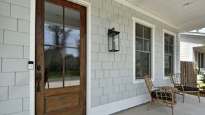 A house with a wooden front door, a porch with two wicker chairs, and a wall-mounted lantern. - Beautiful 4 bedroom home with a pool! (Saint Simons Island)