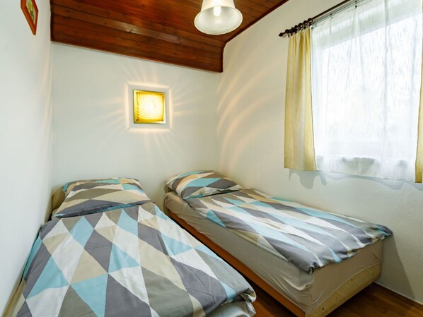 Pequeña habitación con dos camas, techo de madera, luz empotrada en la pared y una ventana con cortinas.