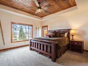 A bedroom with a wooden ceiling, a large window, a bed with a headboard, and a nightstand with a lamp.
