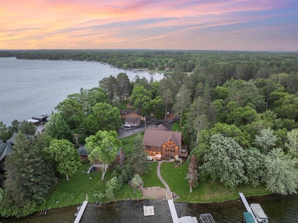 An aerial view of a lakeside property with a large house surrounded by dense trees, a dock, and a boat.