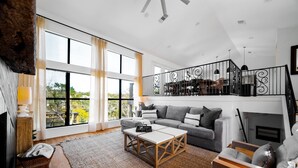A spacious living room with a large window, a grey sofa, a wooden coffee table, and a staircase with a railing.