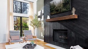 A modern living room with a fireplace, two grey armchairs, a coffee table, and a large window.