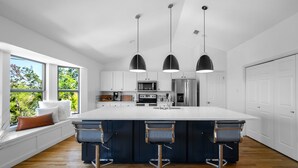 A modern kitchen with a large island, bar stools, pendant lights, and a view of greenery outside.