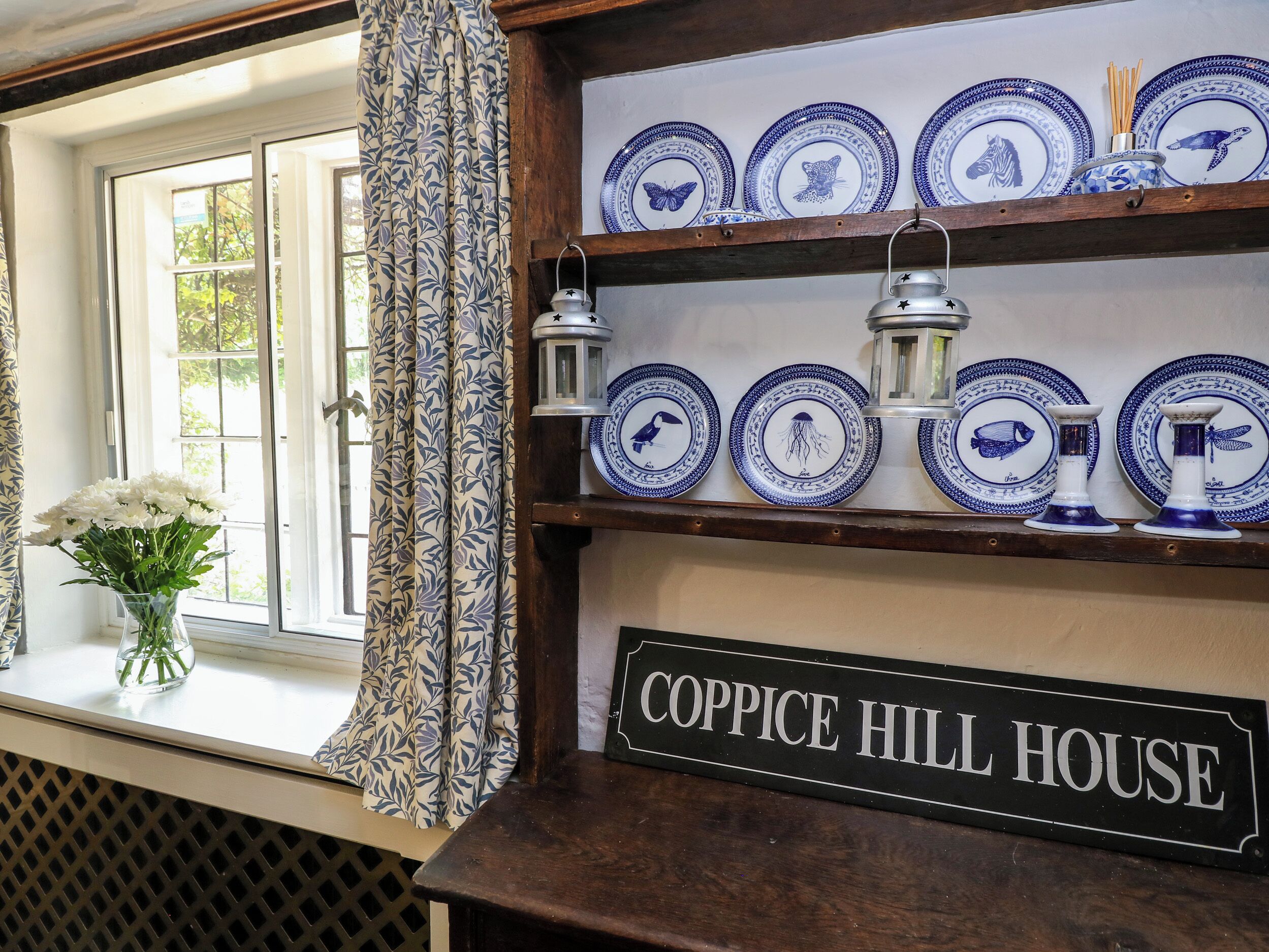 A window with a floral curtain, a vase of white flowers, and a sign reading "COPPICE HILL HOUSE".
