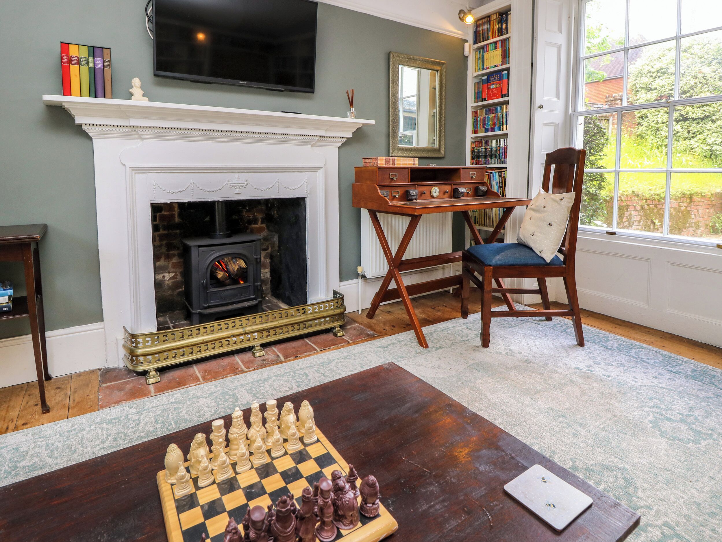 A living room with a fireplace, a chessboard, a bookshelf, and a window with a view of greenery.