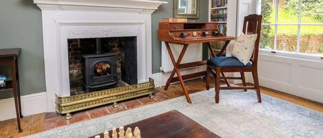 A living room with a fireplace, a chessboard, a bookshelf, and a window with a view of greenery.