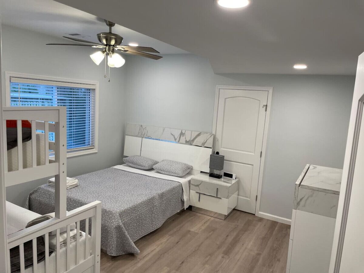Quarto com cama, mesas de cabeceira, ventilador de teto, janela com persianas e porta.