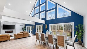 A spacious living area with a high ceiling, large windows, a fireplace, and a dining area with chairs and a table.