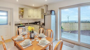 Un coin repas avec une table dressée pour quatre personnes, une cuisine avec des meubles blancs, un réfrigérateur et une vue à travers une porte vitrée.