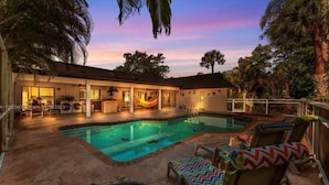 Un área de piscina con sillones de descanso y una hamaca, rodeada de palmeras y una casa con patio.