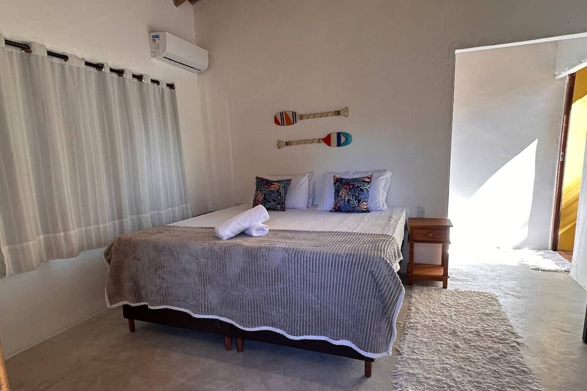 A bedroom with a large bed, a wooden nightstand, a wall-mounted air conditioner, and a window with curtains.