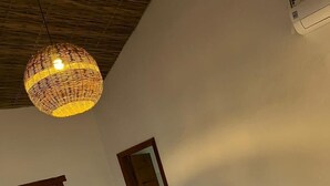 A bedroom with a bed, white pillows, a woven light fixture, a wooden door, and a view of greenery outside. - Villa Beija Flôr (Porto Seguro)