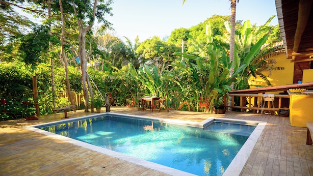 A rectangular swimming pool with a wooden deck, surrounded by lush greenery and a thatched roof structure. - Villa Beija Flôr (Porto Seguro)