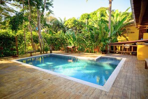 Una piscina rectangular con cubierta de madera, rodeada de abundante vegetación y una estructura de techo de paja.