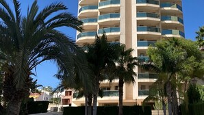 Un hotel alto y moderno con un elemento vertical central, rodeado de palmeras y una piscina.