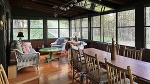 A spacious room with a wooden dining table, wicker chairs, and a green round table. Large windows provide views of the outdoors. - LAKE GARFIELD WATERFRONT HOME AND BOATHOUSE (Monterey)