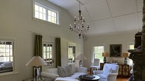 A spacious living room with a white sofa, armchairs, a coffee table, and a chandelier. There are windows with curtains, a painting on the wall, and a stone fireplace. - LAKE GARFIELD WATERFRONT HOME AND BOATHOUSE (Monterey)