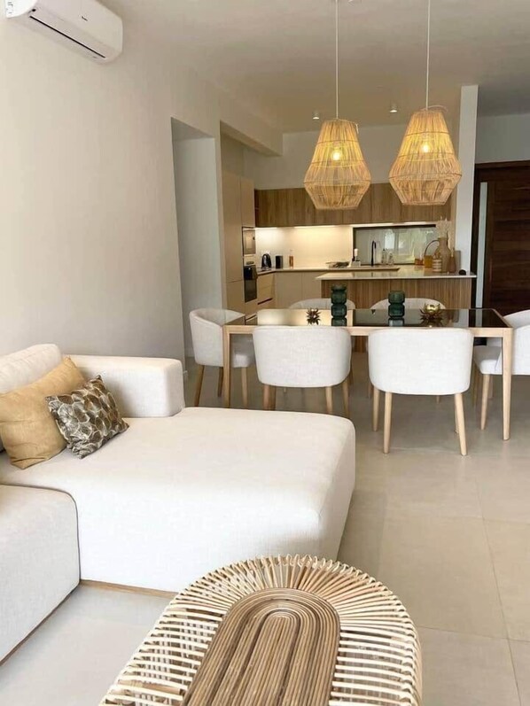 A modern living room with a sofa, dining table, and chairs. There is a kitchen in the background with wooden cabinets and a hanging light fixture.