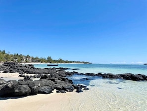 Una playa con agua cristalina, rocas negras y una costa lejana con edificios.