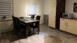 A compact kitchen with a dining table, chairs, a microwave, cabinets, and a window with blinds. - Appartement für 2 Personen in Ruhiger Lage (Ilsede)