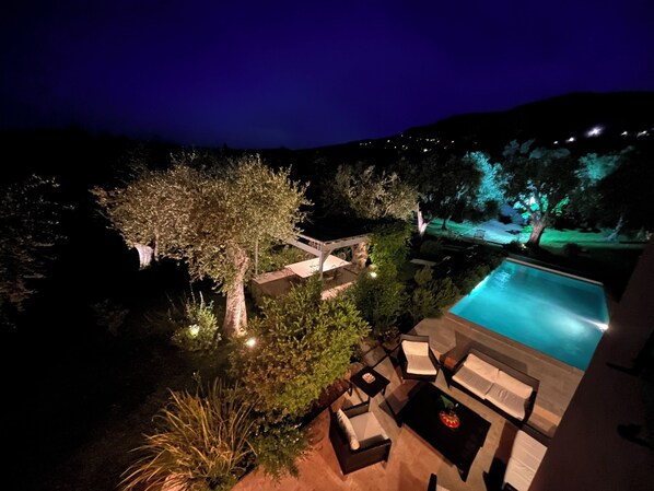 A nighttime view of a pool area with outdoor furniture and a tree.