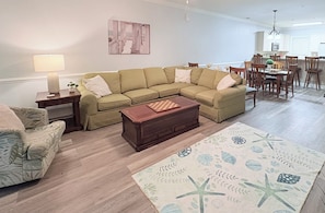 Una sala de estar con sofá modular, una mesa de centro de madera, alfombra estampada y una zona de comedor con sillas y mesa de madera.