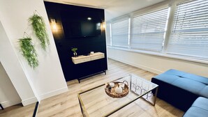 Uma sala de estar moderna com um painel de entretenimento preto, uma mesa de centro de vidro, um sofá azul e uma janela grande com persianas.