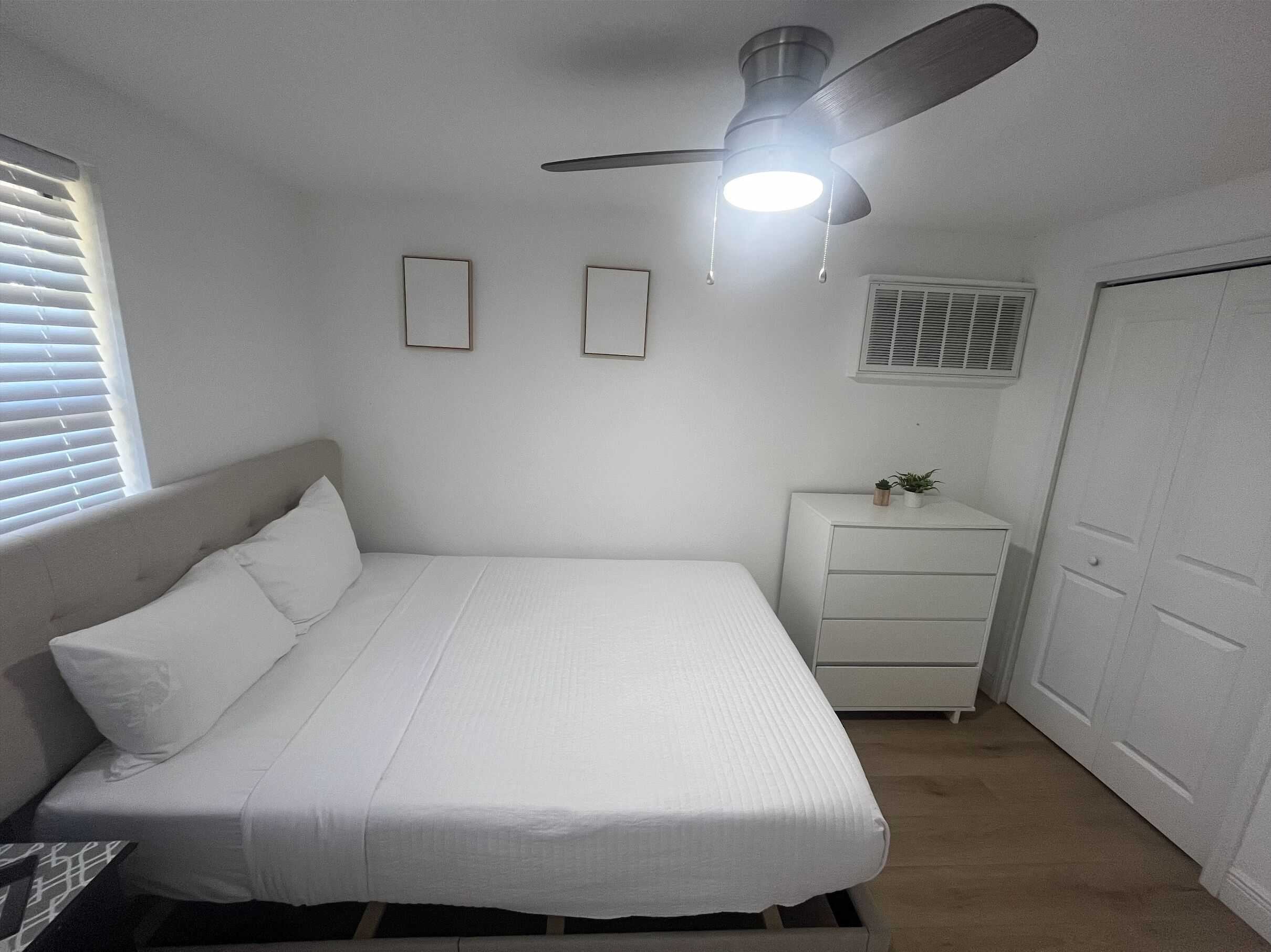 Een slaapkamer met een bed, nachtkastje, plafondventilator en een raam met zonwering.