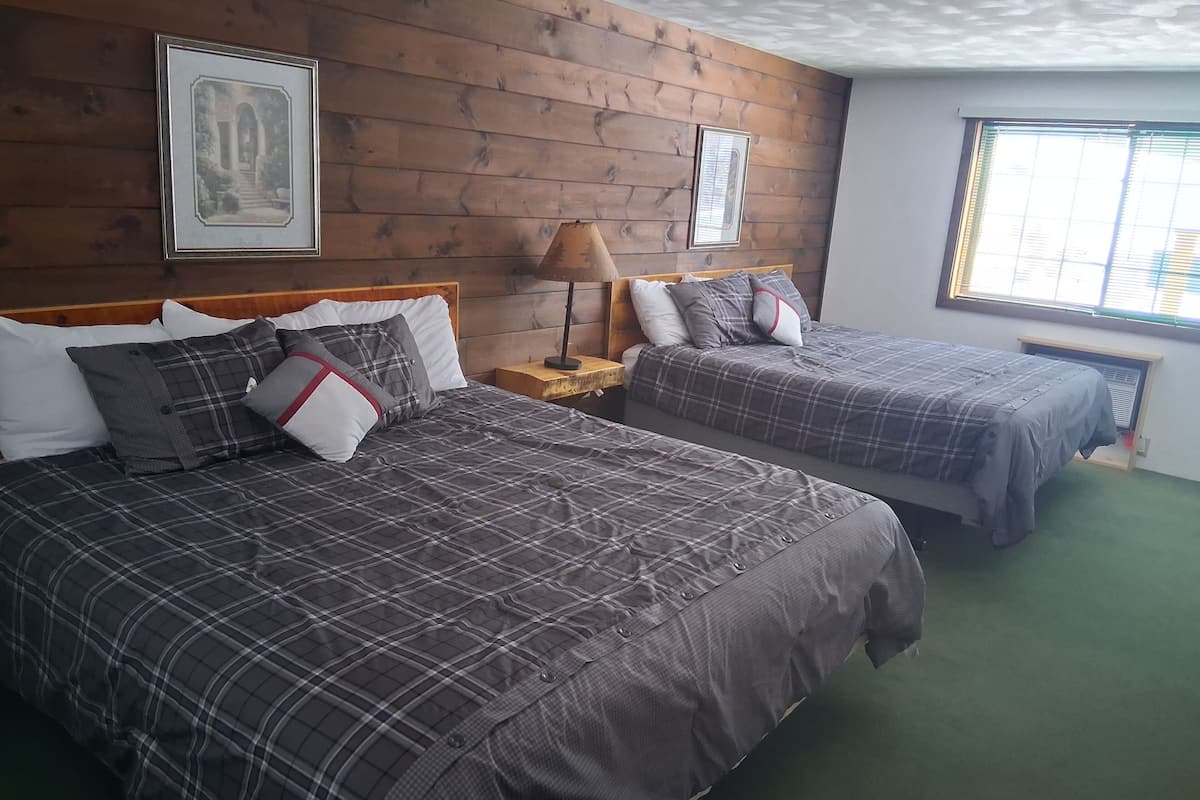 A hotel room with two beds, wooden panel walls, a framed picture, a nightstand, a lamp, and a window with blinds.