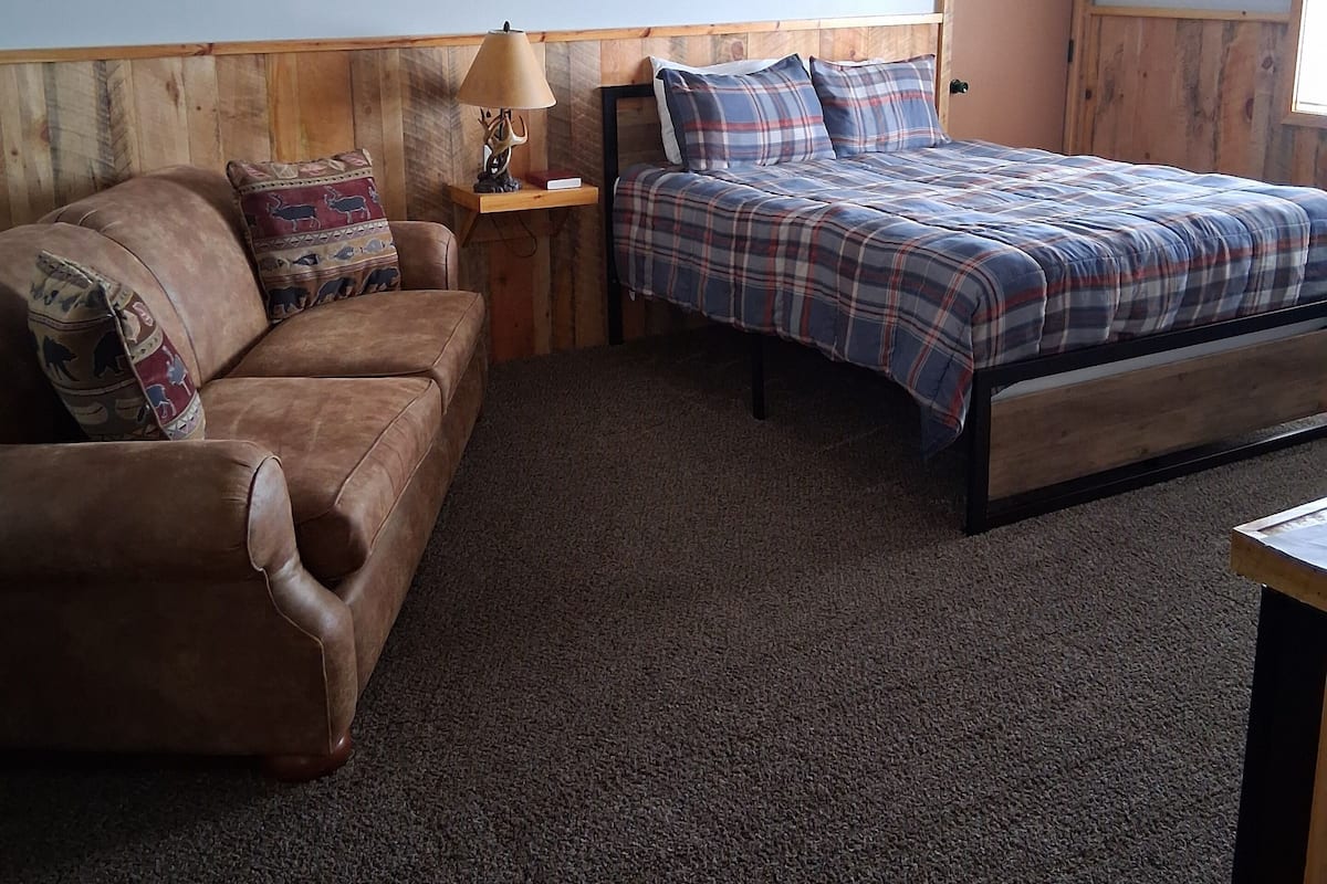 A bedroom with a wooden headboard, a plaid bedspread, a brown leather sofa, and a small table with a lamp.
