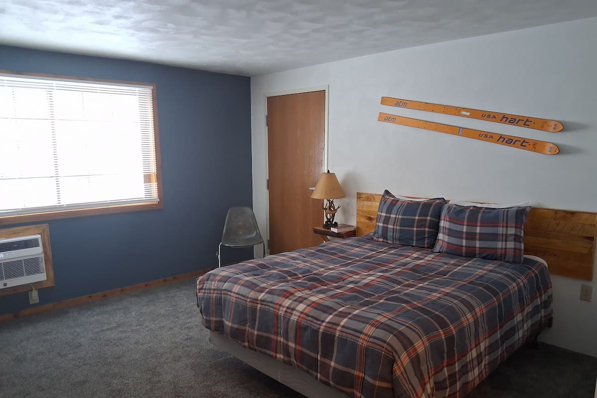 A bedroom with a plaid bedspread, a wooden headboard, a nightstand with a lamp, and a window with blinds.