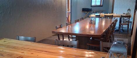 A rustic dining area with a large wooden table and chairs, pendant lighting, and a television mounted on the wall.