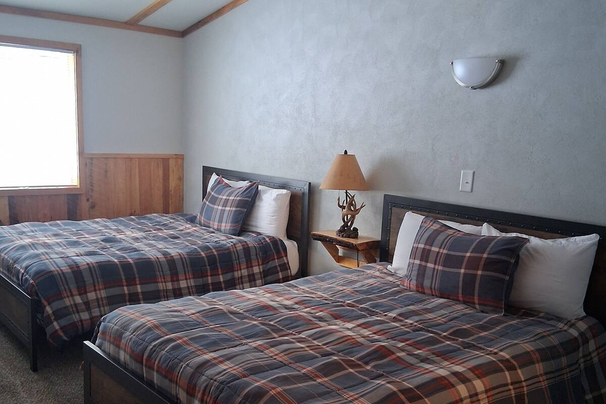A bedroom with two beds, wooden headboards, a nightstand with a lamp, and a window providing natural light.