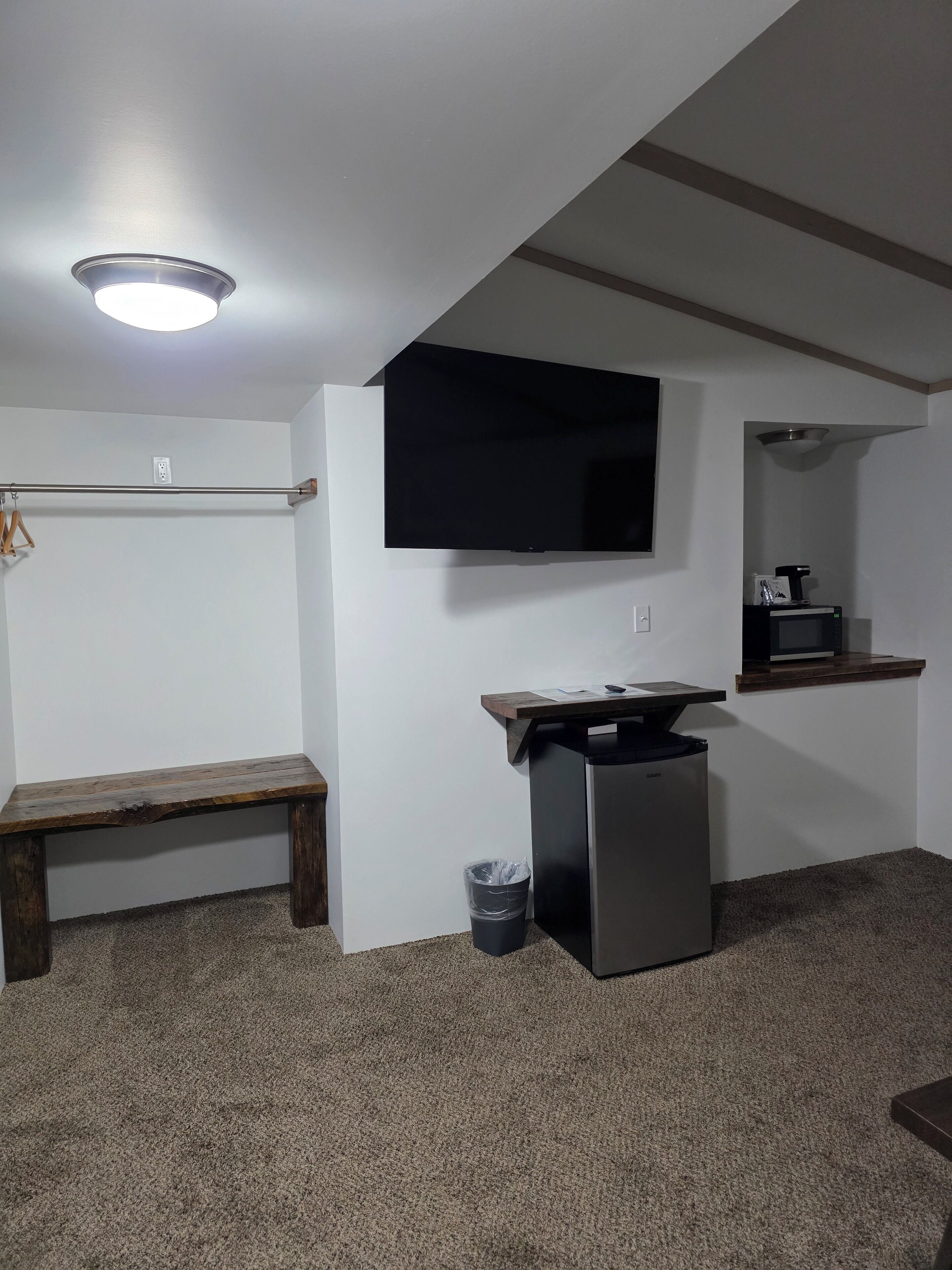 Una habitación pequeña y poco amoblada con una televisión de pantalla plana montada en la pared, una mesita con microondas, un banco y un cubo de basura.