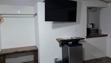 Una habitación pequeña y poco amoblada con una televisión de pantalla plana montada en la pared, una mesita con microondas, un banco y un cubo de basura.