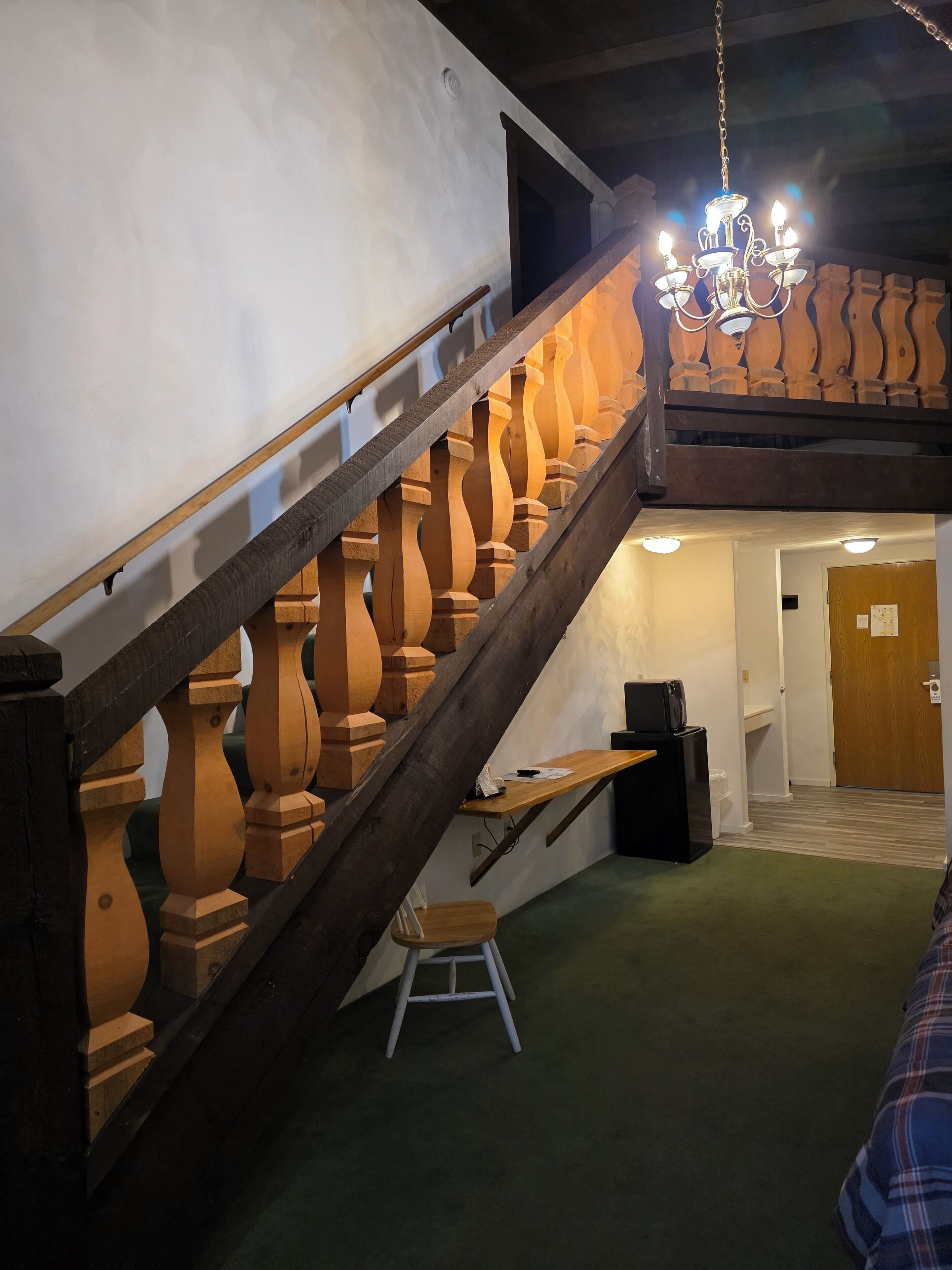 Un escalier avec des balustrades en bois et un lustre, menant à une pièce dotée d’une moquette verte et d’un petit tabouret.