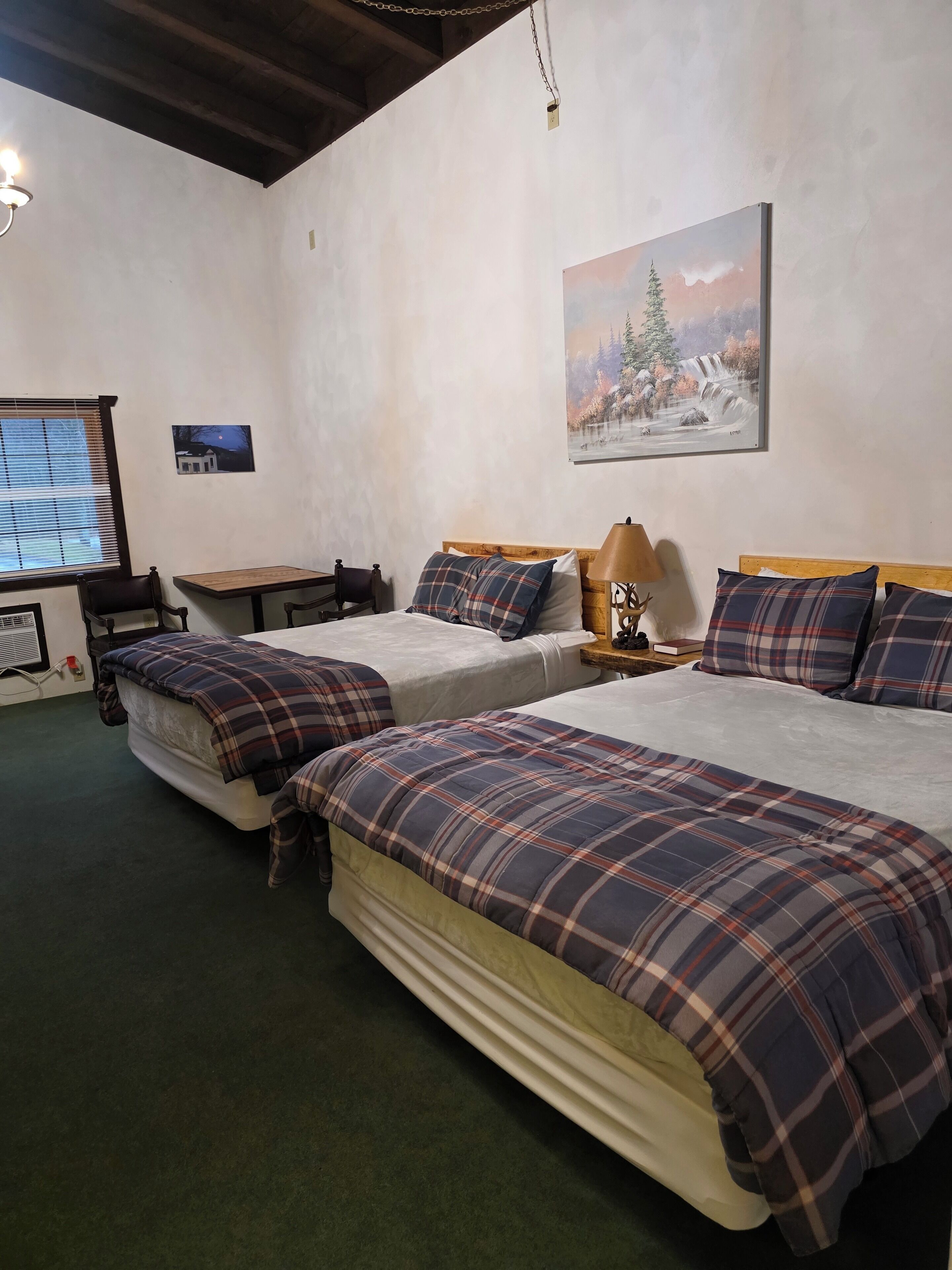 A room with two beds, a table, a chair, a lamp, and a painting on the wall.