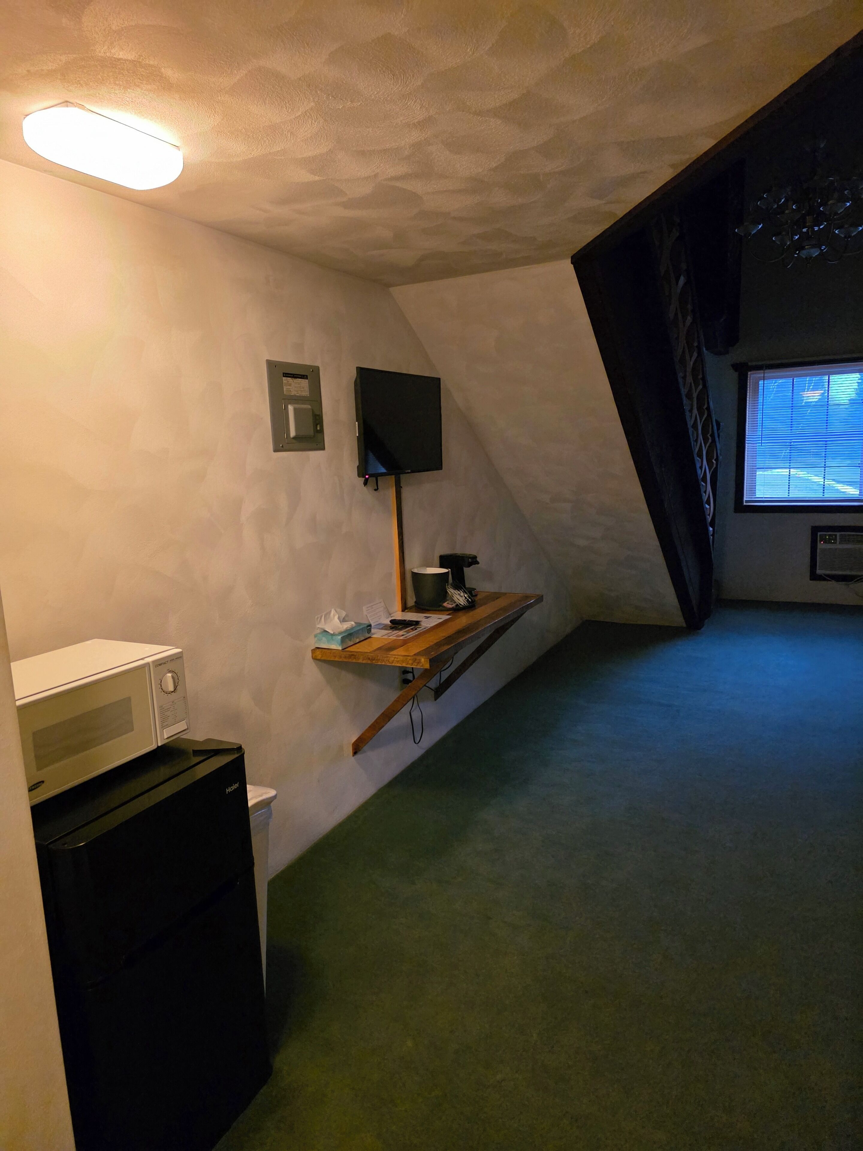Una habitación pequeña y poco iluminada con un microondas, una mesita con una tetera y un televisor montado en la pared.