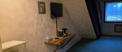 Una habitación pequeña y poco iluminada con un microondas, una mesita con una tetera y un televisor montado en la pared.