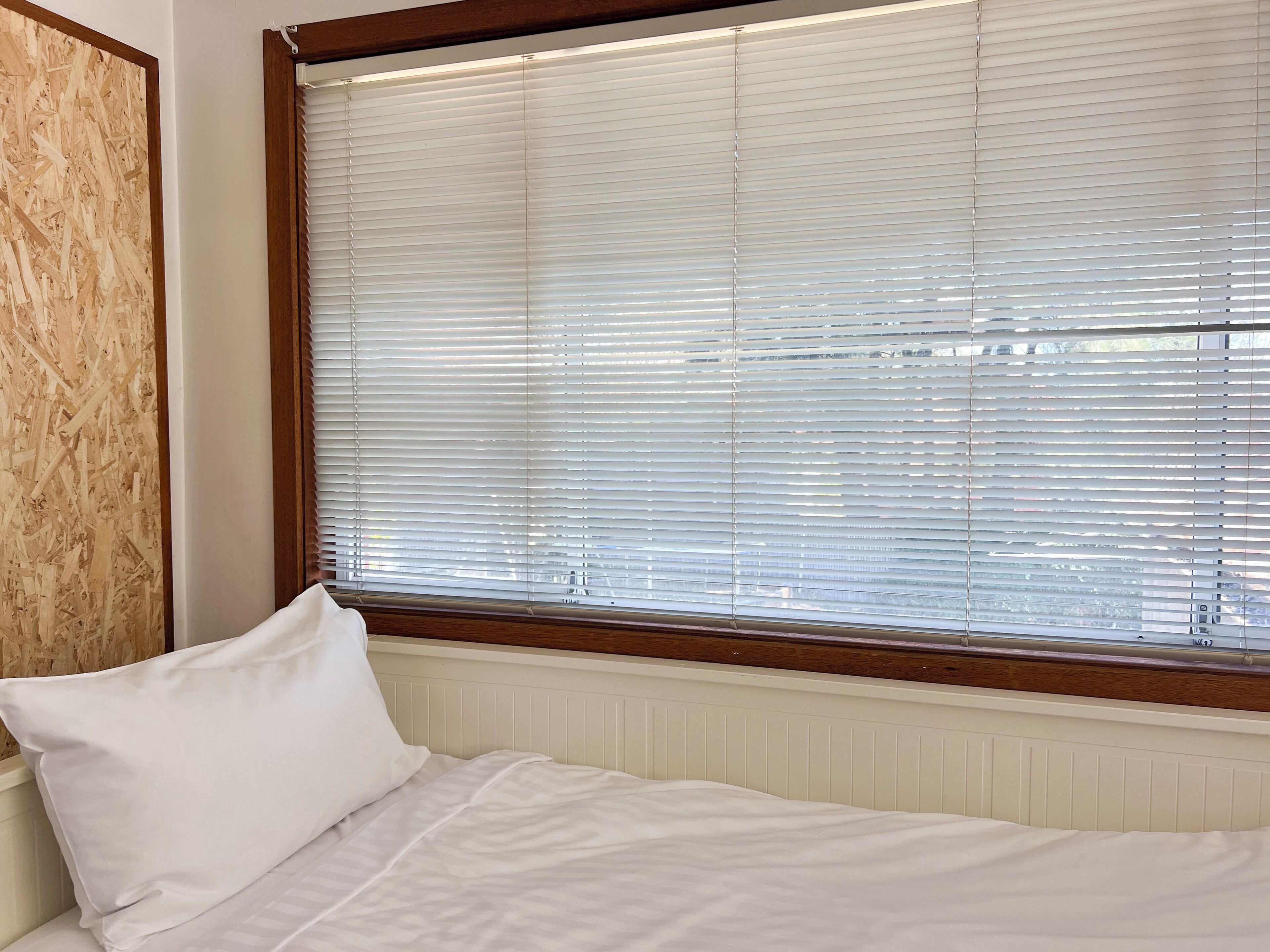 Een slaapkamer met een bed, witte beddengoed, een raam met zonwering en een houten hoofdbord.