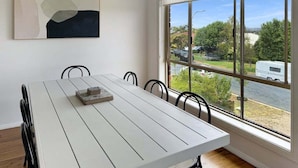 Un coin repas avec une table et des chaises blanches, un tableau au mur et une grande fenêtre donnant sur un quartier résidentiel.