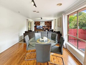 A modern dining area with a glass table surrounded by chairs, a brick accent wall, and a view of a red fence through large windows.