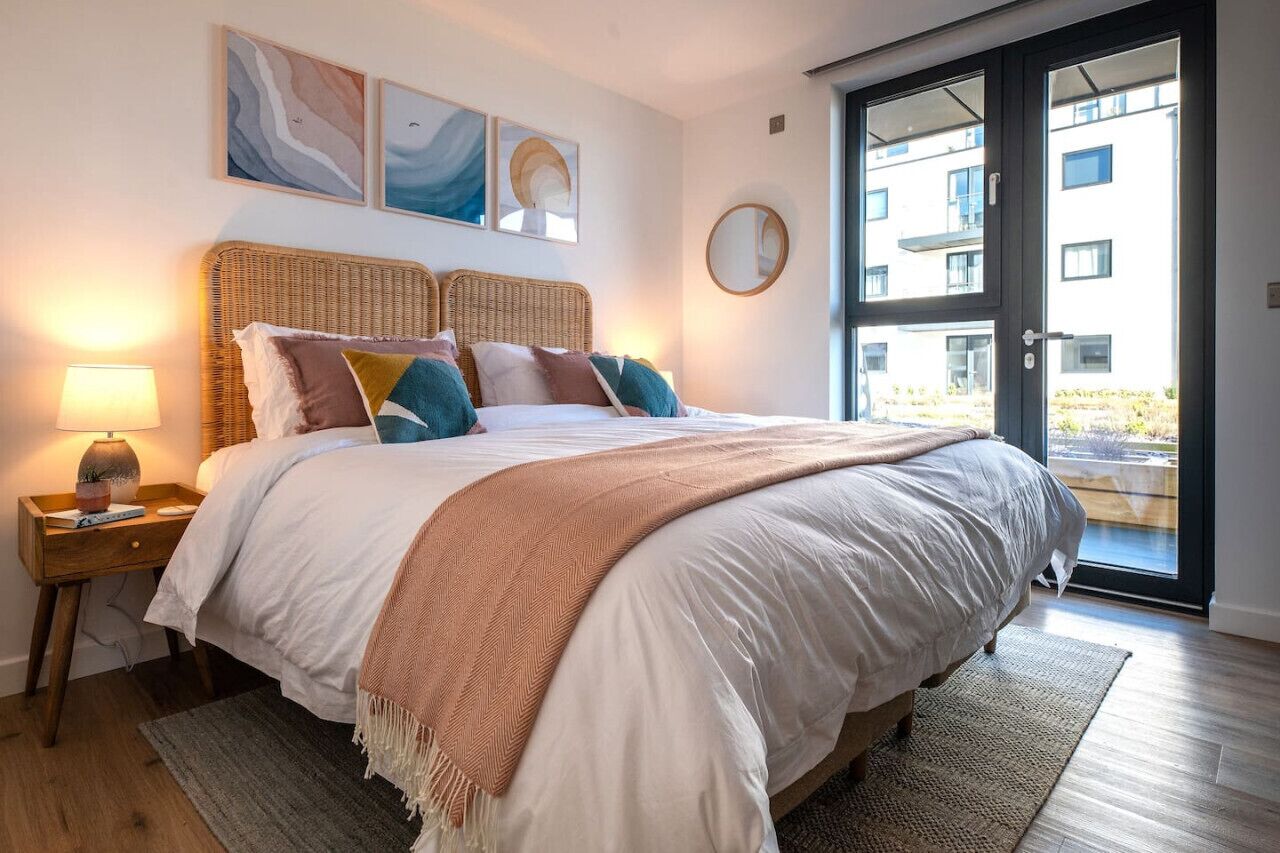 A bedroom with a large bed, a nightstand with a lamp, and a view of an outdoor balcony through a glass door.