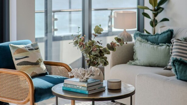 Ein modernes Wohnzimmer mit Meerblick, ausgestattet mit einem blauen Sofa, einem runden Couchtisch mit Büchern und einer Vase sowie einem weißen Sofa mit türkisfarbenen Kissen.